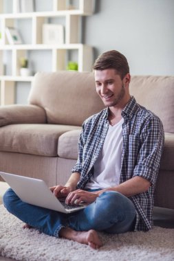 Yakışıklı olan bir dizüstü bilgisayar kullanarak ve yerde evde otururken gülümseyen