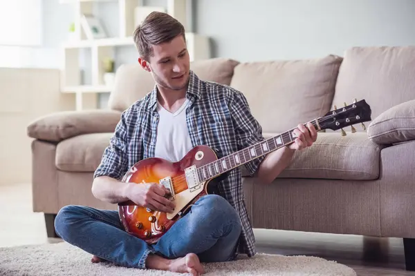 Handsome guy is playing guitar and smiling while sitting on the floor at home