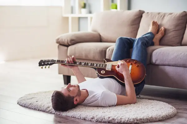 Handsome guy is playing guitar while lying on the floor with his legs on couch