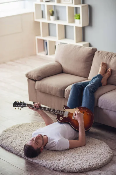Handsome guy is playing guitar while lying on the floor with his legs on couch