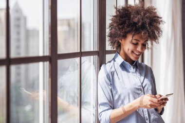 Güzel kız rahat kıyafetler içinde bir akıllı telefon kullanarak ve pencerenin yanında dururken gülümseyen