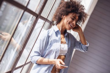 Gündelik giysiler içinde güzel bir kız müzik dinliyor, akıllı bir telefon kullanıyor ve gülümsüyor.