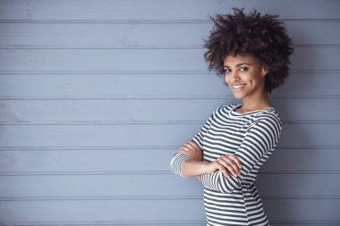 Tarz sahibi kız kameraya bakıp gülümserken kolları çapraz duran gri duvar arka planında duruyor.