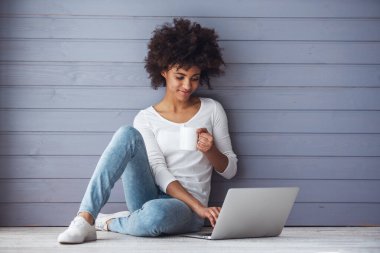 Çekici kız dizüstü bilgisayar kullanıyor, elinde bir bardak tutuyor ve yerde otururken gülümsüyor.