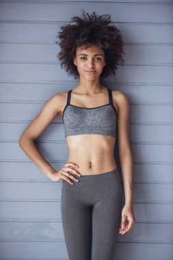 Spor giyim bölümündeki çekici kız gri duvar arkasında kameraya bakıyor ve gülümsüyor.