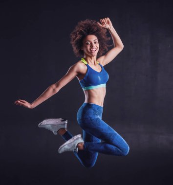 Spor giyinen çekici kız zıplıyor, kameraya bakıyor ve gülümsüyor, siyah arka planda.