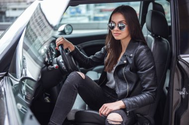 Deri ceket ve güneş gözlüklü güzel kız arabasında oturuyor