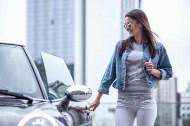 Jean ceket ve güneş gözlüklü güzel kız arabasını açma ve gülümseyen