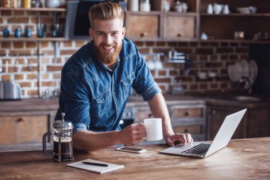 Sakallı yakışıklı genç kahve içme, kameraya bakıyor ve mutfakta bir dizüstü bilgisayar ile çalışırken gülümseyen