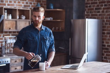 Sakallı yakışıklı genç kahve içme ve mutfakta bir dizüstü bilgisayar ile çalışırken kameraya bakıyor