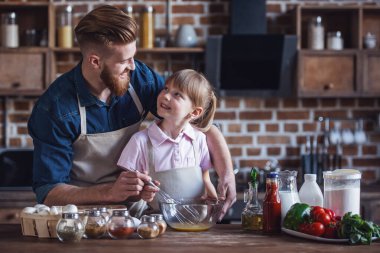 Sevimli küçük kız ve yakışıklı sakallı babası önlük içinde yumurta çırpma ve mutfakta yemek yaparken gülümseyen