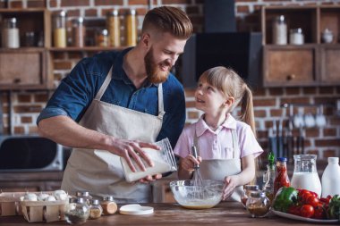 Sevimli küçük kız ve yakışıklı sakallı babası önlük içinde yumurta çırpma, un ekleyerek ve mutfakta yemek yaparken gülümseyen