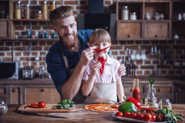 Sevimli küçük kız ve yakışıklı sakallı babası önlük içinde malzemelerle pizza mutfakta yemek yaparken eğleniyor musun