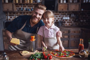 Sevimli küçük kız ve yakışıklı sakallı babası önlük içinde kameraya bakarak ve pizza mutfakta yemek yaparken gülümseyen