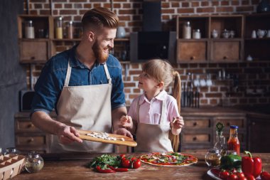 Sevimli küçük kız ve yakışıklı sakallı babası önlük içinde söz ve pizza mutfakta yemek yaparken gülümseyen