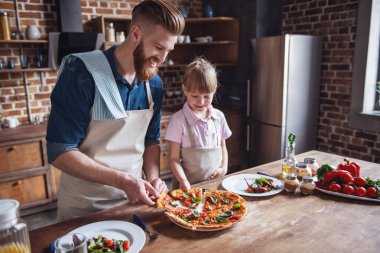 Sevimli küçük kız ve yakışıklı sakallı babası önlük içinde dilim sadece pişmiş pizza atıyor ve gülümseyen