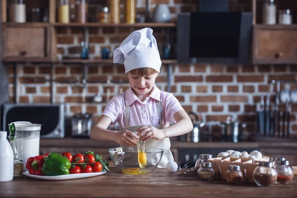 Cute little girl in chef\'s hat and apron is cracking eggs and smiling while cooking in kitchen
