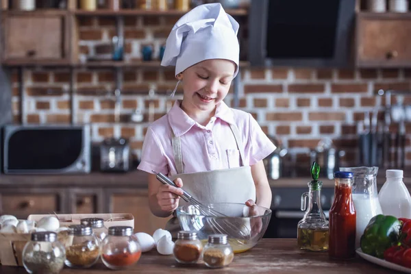 Cute little girl in chef\'s hat and apron is whisking eggs and smiling while cooking in kitchen