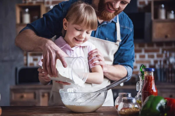 Cute little girl and her handsome bearded dad in aprons are preparing dough, adding flour and smiling while cooking in kitchen