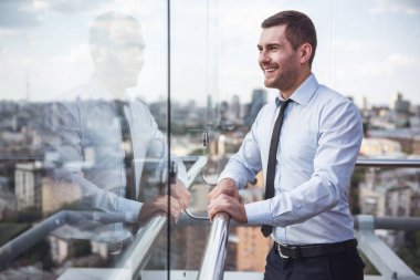 Yakışıklı işadamı pencereden dışarı bakarak ve ofis binası balkonda istirahat ederken gülümseyen