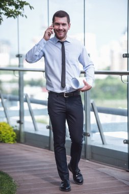 Mobil telefonda konuşurken, haddelenmiş-kağıdı tutan ve açık havada yürürken gülümseyen yakışıklı işadamı portresi uzunluğu tam