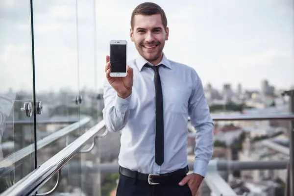 Handsome businessman is showing a smart phone, looking at camera and smiling while standing on balcony of the office building