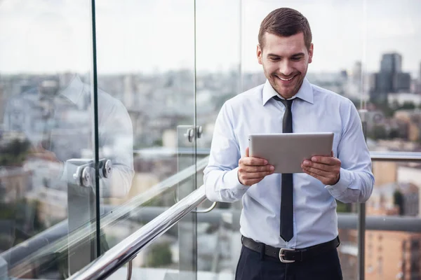 Handsome businessman is using a digital tablet and smiling while resting on balcony of the office building
