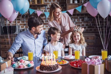 Mutlu aile doğum günü kutlaması sırasında dekore edilmiş mutfak masasında oturuyor, anne kızı üzerinde bir taç koyuyor