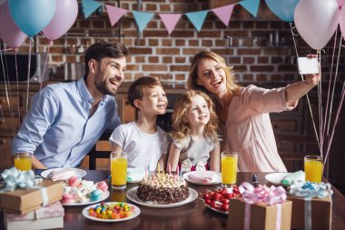 Mutlu bir aile bir akıllı telefon kullanarak ve doğum günü kutlaması sırasında dekore edilmiş mutfak masasında otururken gülümseyen selfie yapıyor