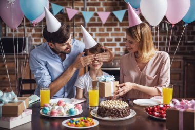 Anne hediye tutarken mutlu aile oturuyor doğum günü kutlaması sırasında dekore edilmiş mutfak masasında baba oğlunun gözleri koruyor