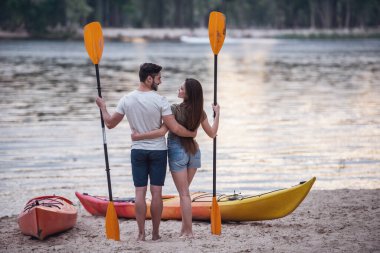 Arkadan görünüşü kürekler tutarak ve süre sahilde kayaks yakınındaki ayakta ve gün batımını izlemek sarılma genç Çift