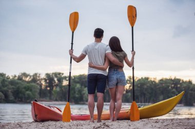 Arkadan görünüşü kürekler tutarak ve süre sahilde kayaks yakınındaki ayakta ve gün batımını izlemek sarılma genç Çift