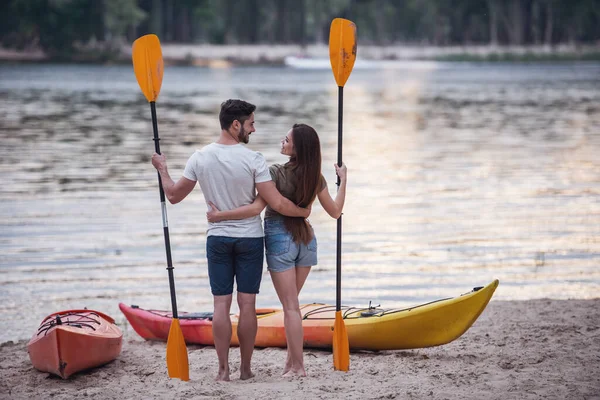 后视图的年轻夫妇抱着桨和拥抱时站在海滩上小艇附近 看着日落 — 图库照片