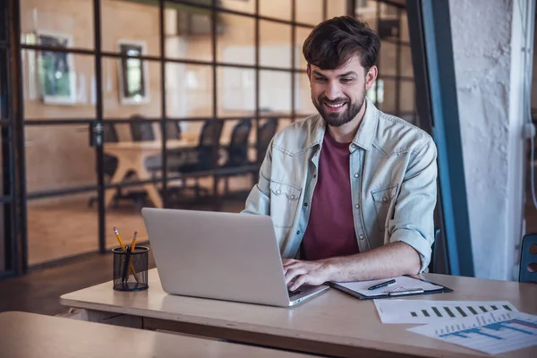 Handsome bearded businessman in casual clothes is using a laptop and smiling while working in office