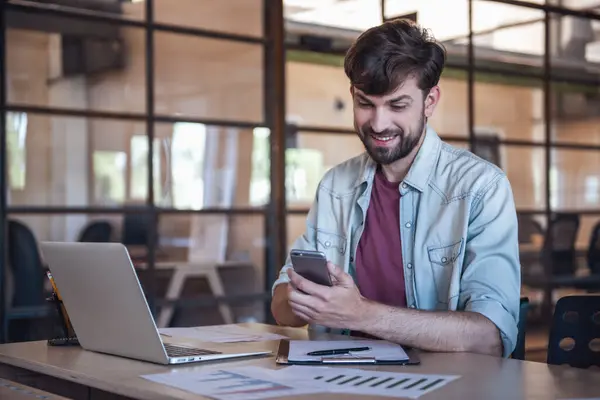 Yakışıklı sakallı iş adamı rahat kıyafetler bir akıllı telefon kullanarak ve bir dizüstü bilgisayar Office ile çalışırken gülümseyen