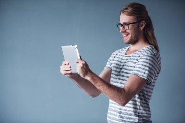 Omzuna ile şık genç adam sarı saçlı ve eyglasses dijital tablet kullanma ve gri arka plan üzerinde gülümseyen