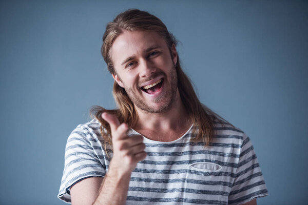 Stylish young man with shoulder-length blond hair is pointing at camera and smiling, on gray background