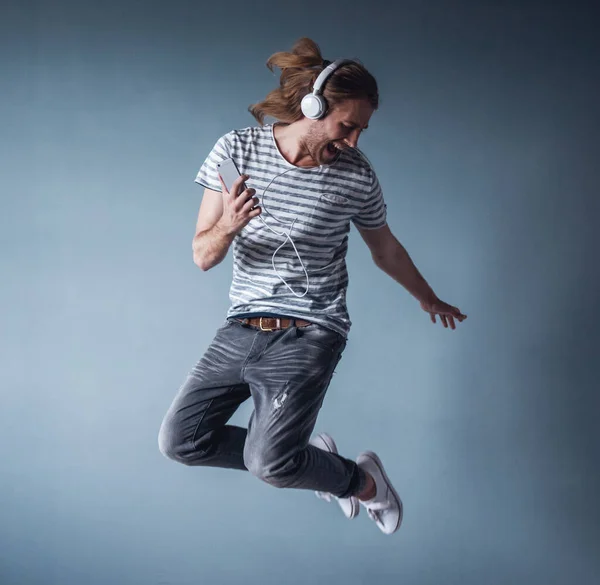 Stylish young man with shoulder-length blond hair and in headphones is listening to music while jumping, on gray background
