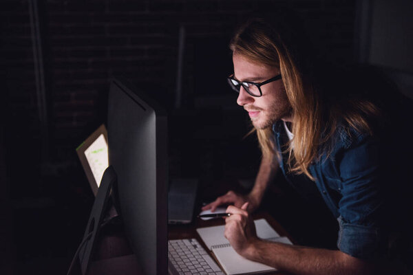 Handsome young businessman with shoulder-length blond hair and in casual wear and eyeglasses is making notes while working with a computer at night