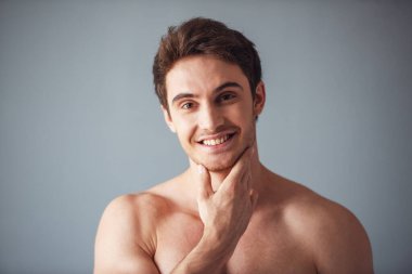 Yakışıklı genç adam onun çene dokunmadan, kameraya bakıyor ve gülümsüyor, gri arka plan üzerinde çıplak gövde ile portresi