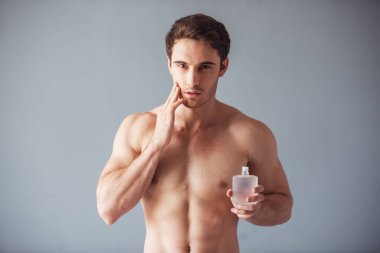 Yakışıklı genç çıplak gövde ile tıraş losyonu holding, çenesine dokunuyor ve seductively gri arka plan üzerinde kamera bakıyor