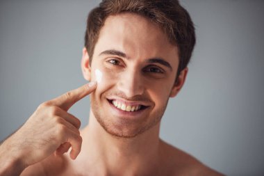 Yakışıklı genç çıplak gövde ile krem yüzünde, kameraya bakıyor ve gülümsüyor, gri arka plan üzerinde uygulanıyor