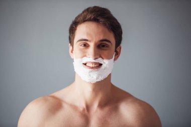 Yakışıklı delikanlı yüzünü tıraş köpüğü ile kameraya bakıyor ve gülümsüyor, gri arka plan üzerinde