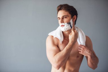Onun yüzünde köpükle yakışıklı genç gri arka plan üzerinde bir jilet kullanarak tıraş oluyor