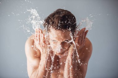 Yakışıklı genç çıplak gövde ile yüzünü etrafında, gri arka plan su sıçramalarına yıkıyor