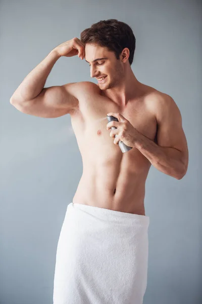 Handsome young man wrapped in bath towel and with bare torso is using a spray deodorant and smiling, on gray background