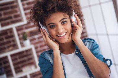 Kulaklıklı güzel kız müzik dinliyor, kameraya bakıyor ve evde vakit geçirirken gülümsüyor.