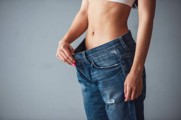 Cropped image of beautiful girl in large size jeans showing her weight loss, on gray background