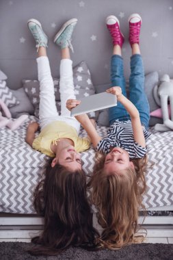 İki sevimli küçük kız dijital tablet kullanma ve sırtlarını çocuk odası yatakta yatarken gülümseyen