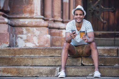 Yakışıklı adam akıllı bir telefon kullanıyor, kahve içiyor ve merdivenlerde otururken gülümsüyor.